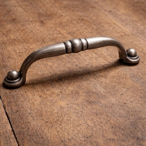 Könnte beinhalten: Ein silberfarbener Schrankgriff mit gebogenem Griff und dekorativen Kugelenden. Die Mitte des Griffs weist drei abgerundete Details auf. Die Hardware befindet sich auf einer warmen Holzoberfläche.