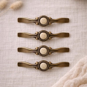 May include: Four antique brass cabinet pulls with a cream-colored center and decorative detailing. The pulls are arranged horizontally on a neutral linen surface, suggesting a vintage or farmhouse aesthetic. Each pull measures approximately 6 inches in length.