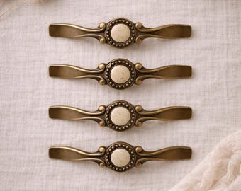 May include: Four antique brass cabinet pulls with a cream-coloured centre and decorative detailing. The pulls are arranged horizontally on a neutral linen surface, suggesting a vintage aesthetic. Each pull measures approximately 15 cm in length.