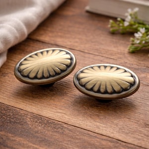 May include: Two antique brass oval cabinet knobs with a floral design. The knobs have a dark base and a detailed, fan-like pattern on the top. They are set on a wooden surface.