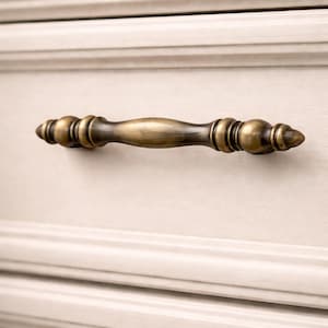 May include: A bronze-toned cabinet pull with a decorative, elongated design. The hardware is mounted on a cream-colored cabinet door, showcasing the pull's detailed, symmetrical form. The pull is approximately 6 inches long.