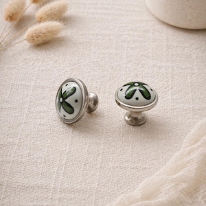 Puede incluir: Dos tiradores decorativos para armarios. Uno es ovalado, el otro redondo. Ambos tienen un fondo blanco con un diseño floral verde y una base de metal plateado. Los tiradores están sobre un fondo neutro.