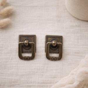 Pode incluir: Duas puxadores de gaveta de bronze antigo com placa traseira retangular e alça decorativa. Os puxadores têm uma estética vintage, com detalhes intrincados e um acabamento quente e envelhecido. São ideais para adicionar um toque de elegância clássica aos móveis.