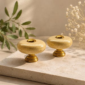 Può includere: Due pomelli decorativi per mobili. Ogni pomello presenta una parte superiore in ceramica beige chiaro macchiettata con un accento e una base in metallo dorato. I pomelli sono esposti su una superficie di pietra colorata.