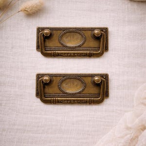 May include: Two antique brass drawer pulls with oval center details and decorative handles. The pulls are rectangular with rounded edges and small ball-shaped accents. They are set against a neutral, textured background.