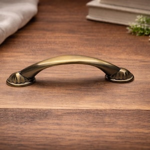 May include: A bronze cabinet pull handle with a curved design. The handle has decorative detailing at each end and is set against a wooden surface. The hardware is likely for furniture or cabinetry.