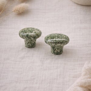 May include: Two mushroom-shaped cabinet knobs. The knobs are a speckled green and white stone. They have a rounded top and a short, cylindrical base. The knobs are set against a neutral background.