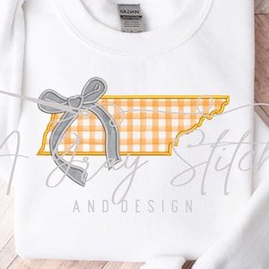 May include: White sweatshirt featuring a design of the state of Tennessee with a yellow and white gingham pattern and a gray bow. The text "A Cozy Stitch AND DESIGN" is printed below the design.
