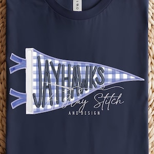 Könnte beinhalten: Marineblaues T-Shirt mit einem Wimpel-Design. Der Wimpel hat ein blau-weißes Vichy-Muster und den Text "JAYHAWKS Day Stitch AND DESIGN" in weißer Schrift. Das T-Shirt wird auf einer gewebten, kreisförmigen Matte präsentiert.