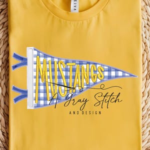 May include: Mustard yellow t-shirt with a pennant design. The pennant is blue and white checkered with the word "MUSTANGS" and the text "A Gray Stitch AND DESIGN". The shirt is folded on a woven, circular surface.