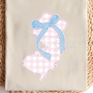 May include: A beige t-shirt with a round neck, featuring an applique of the state of New Jersey in pink gingham, tied with a light blue bow. The shirt is folded and placed on a woven, circular mat.