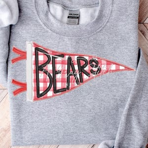 May include: A heather gray sweatshirt with a pennant design. The pennant is red and white checkered with the word "BEARS" in black block letters. Red ribbons are attached to the pennant. The sweatshirt is folded on a wooden surface.