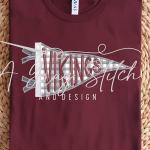 May include: A maroon t-shirt with a crew neck, featuring a graphic of a pennant with the words "Vikings Stitch and Design" in a stylized font. The pennant has a gray and white plaid pattern.