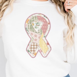 May include: White sweatshirt with a pink and green patchwork ribbon design. The ribbon is made up of different patterns, including floral, polka dot, plaid, and striped.