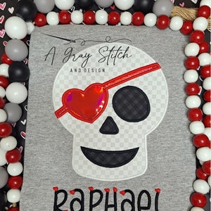 Könnte beinhalten: Ein graues T-Shirt mit einem weißen und schwarzen Totenkopf-Design mit einem roten Herzen als Augenklappe. Der Text "A Gray Stitch and Design" befindet sich über dem Totenkopf und der Name "Raphael" unter dem Totenkopf.