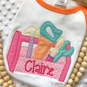 May include: A pink and white toolbox with a saw, hammer, wrench, and screwdriver. The toolbox has a pink polka dot pattern and the name "Claire" is embroidered on the front.