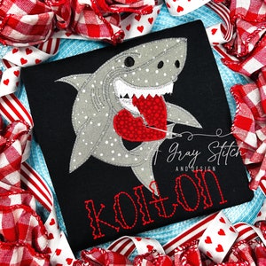 May include: A black t-shirt with a gray shark holding a red heart with white polka dots. The shark has white polka dots and a white mouth. The text "Kolton" is stitched in red on the shirt. The text "Gray Stitch and Design" is stitched in white on the shirt.