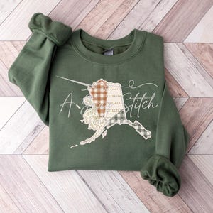 May include: A green sweatshirt with a patchwork design of the state of Alaska and the text "A Stitch" on the front.