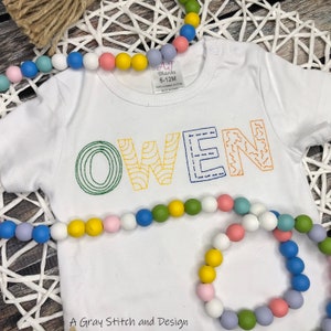May include: White baby onesie with the name "OWEN" embroidered in colorful, patterned letters. The onesie has a tag that says "AJ Blanks 6-12M 100% Combed Cotton Made in China".