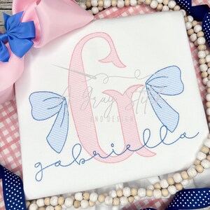 May include: A white t-shirt with a pink embroidered letter G and a blue embroidered bow. The text "gabriella" is embroidered in blue below the letter.
