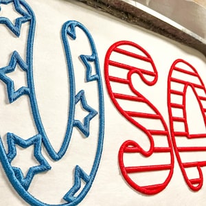 Peut inclure: Broderie "USA" en fil rouge et bleu. Le "U" est bordé d'étoiles bleues et le "S" et le "A" sont rayés de rouge et de blanc.