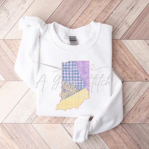 May include: White sweatshirt with a graphic of the state of Indiana in a patchwork design with blue, yellow, and purple colors. The text "A good stitch" is visible on the sweatshirt.