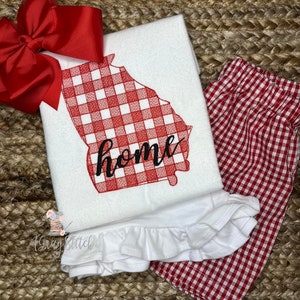 May include: A white shirt with a red gingham Georgia outline and the word "home" in black script. A large red bow is at the top left. Red and white gingham shorts are on the right.