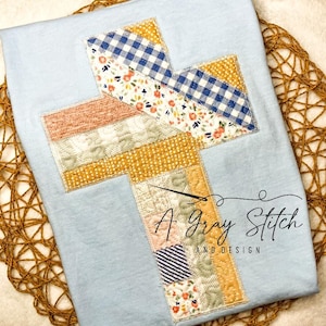 May include: A light blue t-shirt with a patchwork cross design in shades of yellow, pink, and blue. The cross is outlined in gold thread. The text "A Gray Stitch and Design" is printed below the cross.