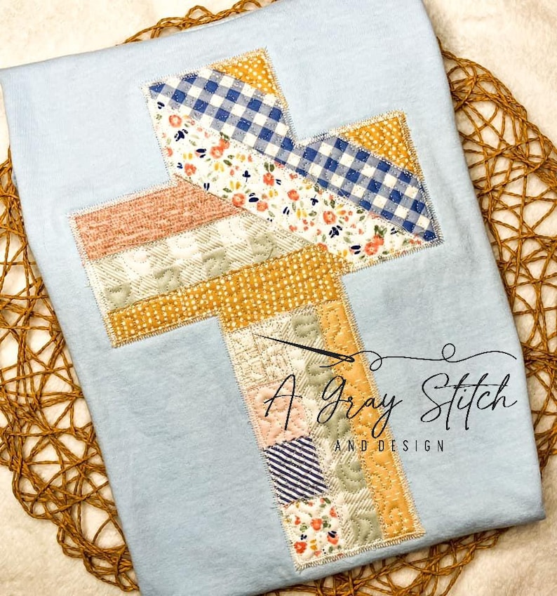 Puede incluir: Una camiseta azul claro con un dise&ntilde;o de cruz de patchwork en tonos de amarillo, rosa y azul. La cruz est&aacute; bordada con hilo dorado. El texto "A Gray Stitch and Design" est&aacute; impreso debajo de la cruz.