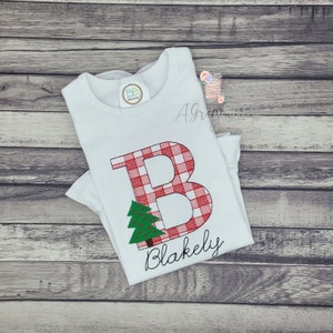 May include: White t-shirt with a red and white plaid letter B and a green Christmas tree. The name "Blakely" is written below the letter.