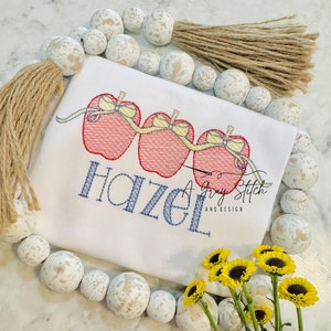 May include: White shirt with a blue embroidered name "Hazel" and three red apples with yellow bows and a yellow ribbon connecting them. The shirt is surrounded by white beads with brown tassels.
