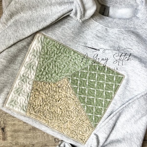 May include: A grey sweatshirt with a square patch of quilted fabric in shades of green and beige. The fabric is stitched in different patterns, including a grid, swirls, and leaves. The text "A Gray Stitch and Design" is embroidered on the patch.
