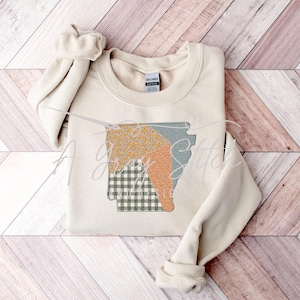 May include: A beige sweatshirt with a patchwork design of a state outline. The design features a green and white plaid pattern, a light orange and yellow pattern, and a light green pattern.