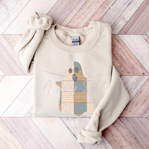 May include: A beige sweatshirt with a patchwork ghost design in shades of yellow, orange, and gray. The ghost has a friendly face and is wearing a plaid pattern.
