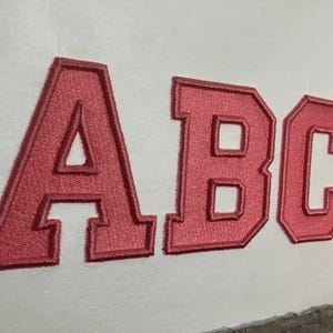 May include: Close-up of the letters "ABC" in a bold, collegiate-style font. The letters are a vibrant red color with a textured, embroidered appearance. The background is a plain white surface.