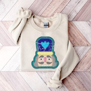 May include: A beige sweatshirt with an embroidered nutcracker design. The nutcracker is wearing a blue hat with a heart on it and has a green beard and mustache.