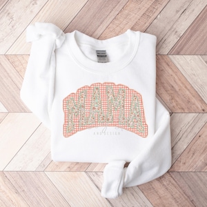 Puede incluir: Sudadera blanca con la palabra "MAMA" en un diseño floral y de cuadros vichy. El texto está arqueado sobre un fondo de cuadros vichy color melocotón. La sudadera también tiene el texto "AND DESIGN" debajo de la palabra "MAMA".