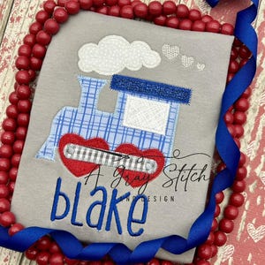 May include: A gray shirt with a blue and white train applique with red hearts. The name "Blake" is embroidered in blue below the train.