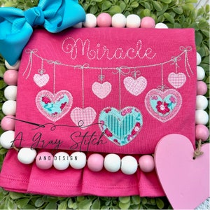 May include: A pink shirt with a white ruffle at the bottom. The shirt has a silver glitter design of a string of hearts with the word "Miracle" above it.