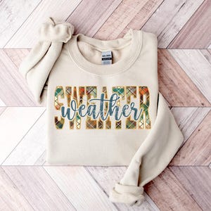 May include: A beige sweatshirt with a plaid pattern design that says "sweater weather" in blue text.
