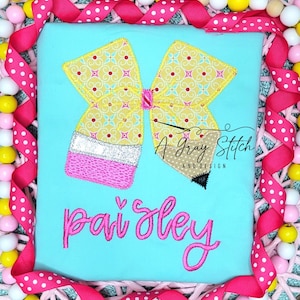 May include: A turquoise blue t-shirt with a yellow and pink bow and pencil design. The text "A Gray Stitch and Design" is in the upper right corner. The name "Paisley" is embroidered in pink below the design.