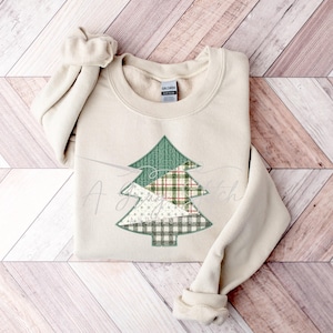 以下が含まれることがあります： ベージュのトレーナーに、緑、白、赤のパッチワークでできたクリスマスツリーのデザインが施されています