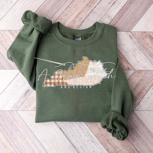 May include: A green sweatshirt with a patchwork design of the state of Kentucky. The design includes a floral pattern, plaid fabric, and a beige background. The text "Stitch and Design" is printed below the state.