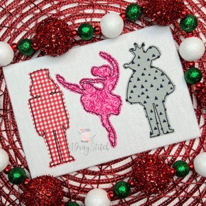 May include: Three Christmas-themed embroidery designs: a nutcracker in red and white gingham, a ballerina in pink with a floral pattern, and a princess in gray with a triangle pattern. The designs are on a white fabric background.