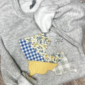 May include: A gray sweatshirt with a patchwork design in the shape of Louisiana. The patchwork is made of blue and white gingham, yellow floral fabric, and a gray and white floral fabric. The sweatshirt has the text "A Gray Stitch" and "Hand Design" on it.