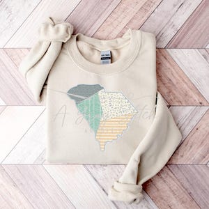 May include: A beige sweatshirt with a patchwork design of the state of South Carolina in green, yellow, and white.