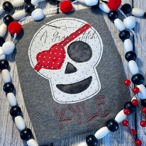 Könnte beinhalten: Ein graues T-Shirt mit einem weißen Totenkopf- und Knochen-Design. Der Schädel hat ein rotes Herz über einem Auge und ein rotes Kreuz über dem anderen Auge. Der Text "A Gray Stitch" befindet sich über dem Schädel und der Name "KYLE" unter dem Schädel.