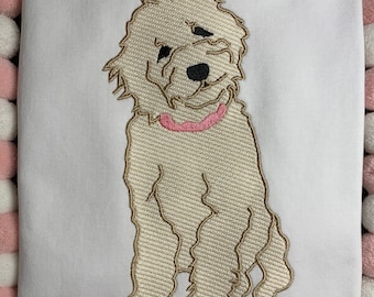 May include: Embroidered design of a seated, fluffy dog with a pink collar on a white background. The dog is beige with black eyes and nose. The embroidery has a textured appearance. The background is surrounded by pink and white pom-poms.