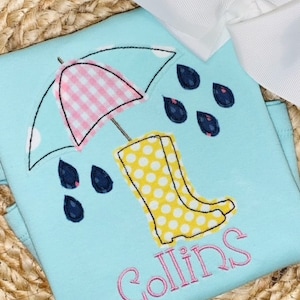 Könnte beinhalten: Hellblaues T-Shirt mit gesticktem Design eines rosa-weiß karierten Regenschirms, gelben gepunkteten Gummistiefeln und marineblauen Regentropfen. Der Name "Collins" ist in rosa Schrift gestickt. Eine weiße Schleife ist im Hintergrund.