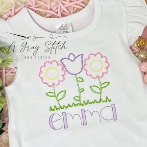 以下が含まれることがあります： 白いベビーロンパースに刺繍された花と紫色の「emma」という名前。花はピンク、黄色、緑です。